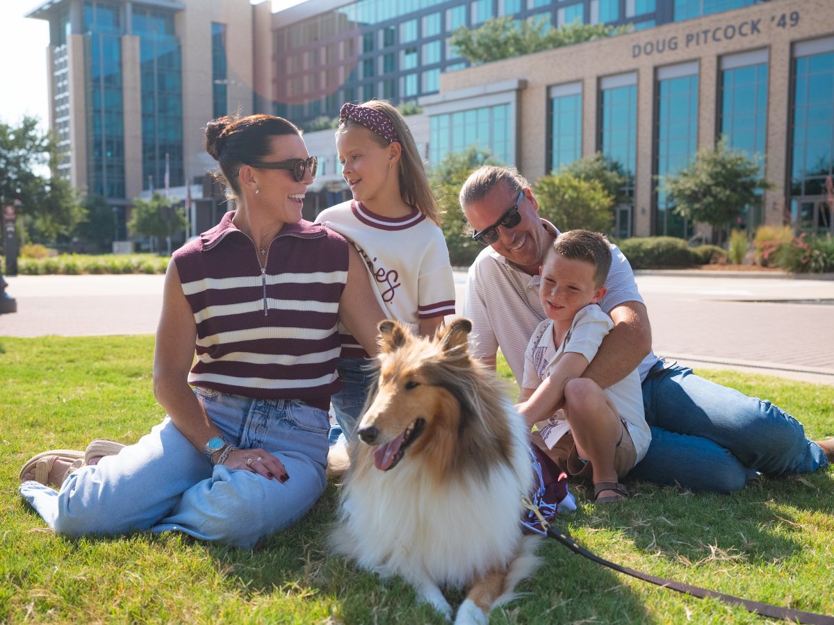A smiling family sits on the grass with a fluffy dog in front, outdoors near a modern building and trees on a sunny day.