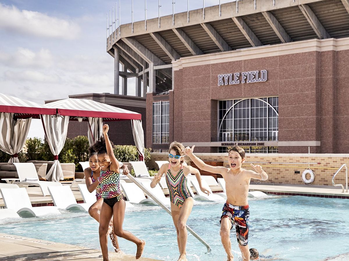 Three kids are jumping into a pool at a hotel water area, with a large stadium-like building in the background, sunny day.