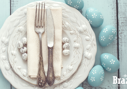 A neatly set plate with a folded napkin, fork, and knife, surrounded by blue Easter eggs on a light wooden table, with "Brazos" watermark.