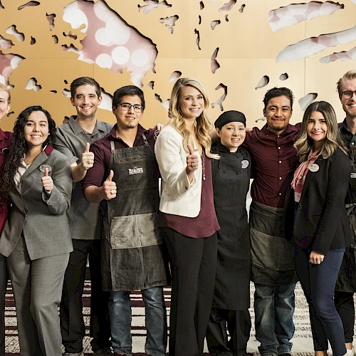 A diverse group of people in aprons posing and giving thumbs up, standing together in a modern indoor setting.