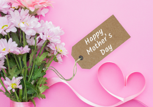 A bouquet of pink and white flowers with a brown tag reading &ldquo;Happy Mother&rsquo;s Day,&rdquo; tied to a pink ribbon on a pink background.