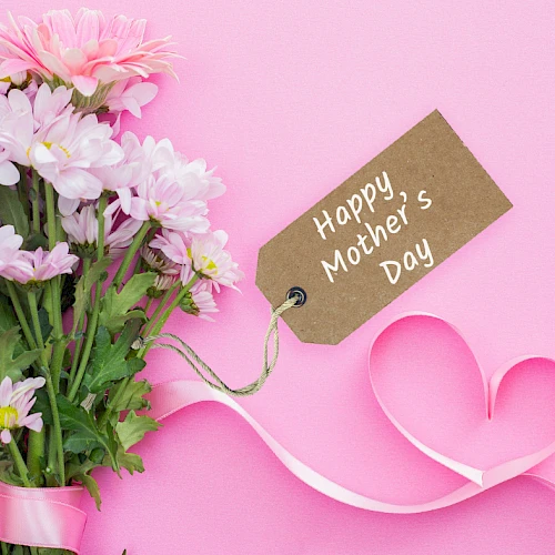 A bouquet of pink and white flowers with a brown tag reading &ldquo;Happy Mother&rsquo;s Day,&rdquo; tied to a pink ribbon on a pink background.