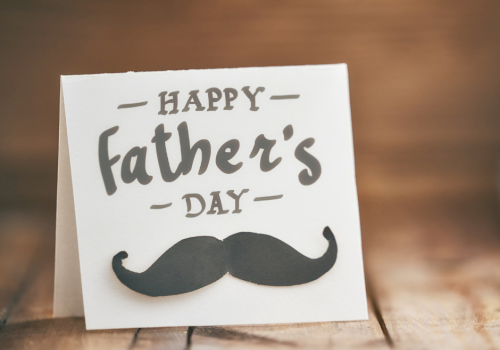 A Father's Day greeting card with a black mustache graphic on a wooden table, warm lighting, and handwritten text.