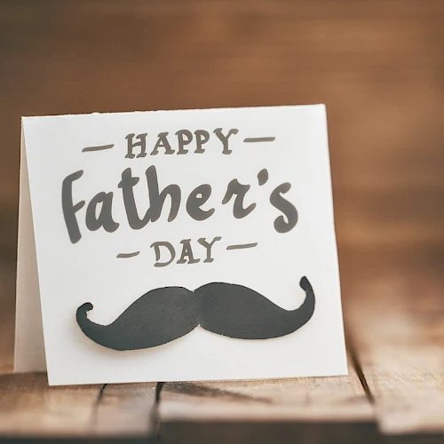 A Father's Day greeting card with a black mustache graphic on a wooden table, warm lighting, and handwritten text.