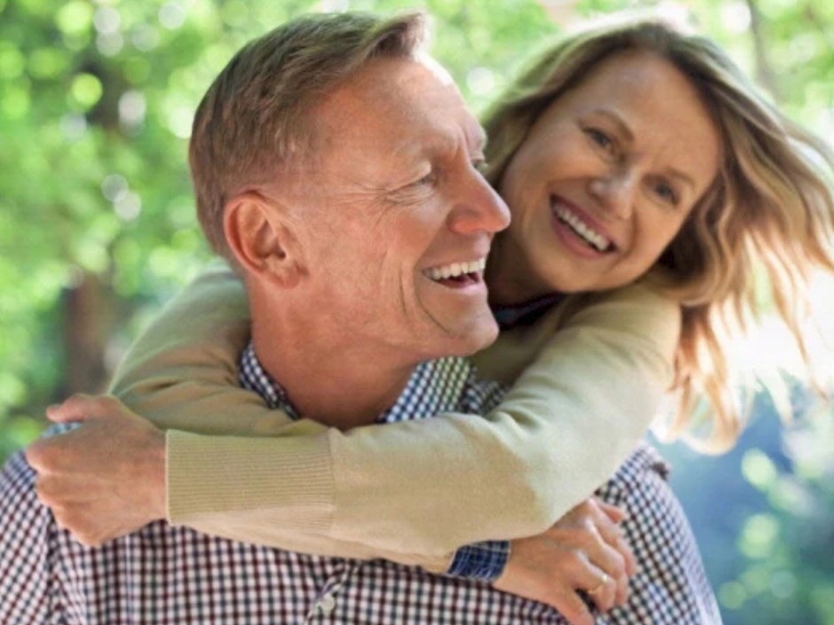 A happy couple outdoors, smiling; a man carries a woman on his back, enjoying a sunny moment together.