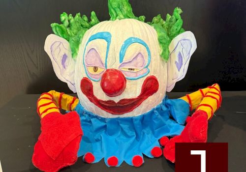 A colorful clown plush with a blue outfit, green hair, red nose, big red lips, yellow sleeves, and red shoes, sitting on a dark surface.