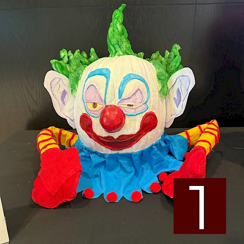 A colorful clown plush with a blue outfit, green hair, red nose, big red lips, yellow sleeves, and red shoes, sitting on a dark surface.