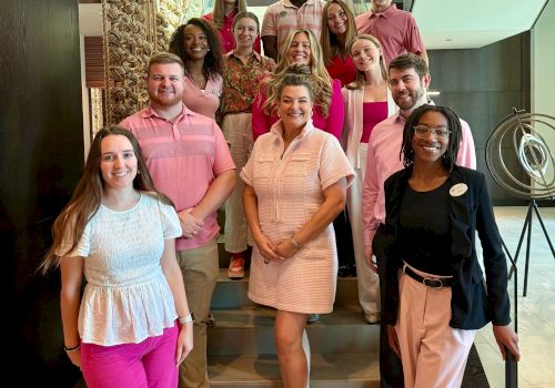 A diverse group of colleagues posing on a staircase inside a modern building. Everyone smiles, casual business attire, posing for a team photo.