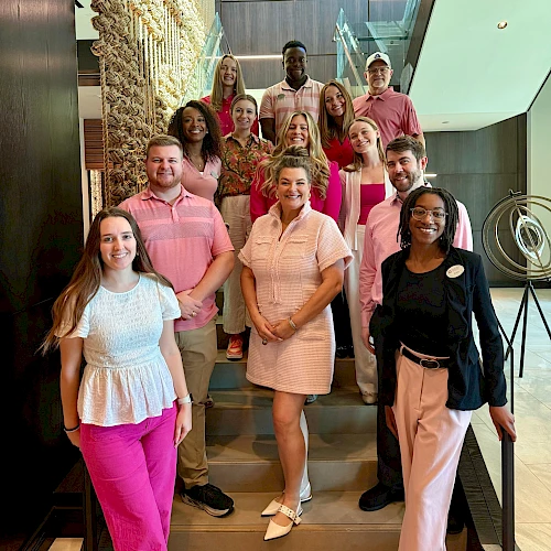 A diverse group of colleagues posing on a staircase inside a modern building. Everyone smiles, casual business attire, posing for a team photo.