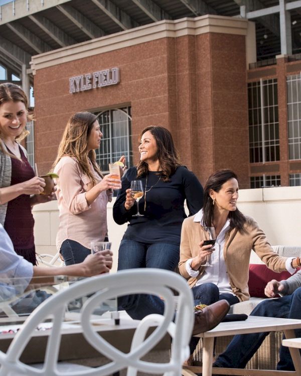 Guests enjoying drinks and conversation around outdoor seating on 2nd floor balcony overlooking Kyle Field