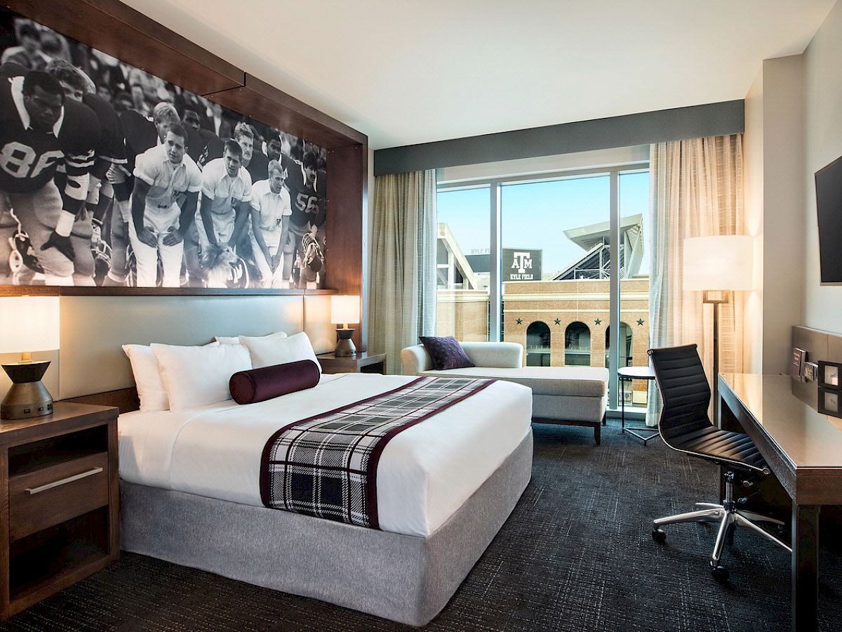 A modern hotel room with a large bed, desk, chair, and a sports-themed black and white photo on the wall.