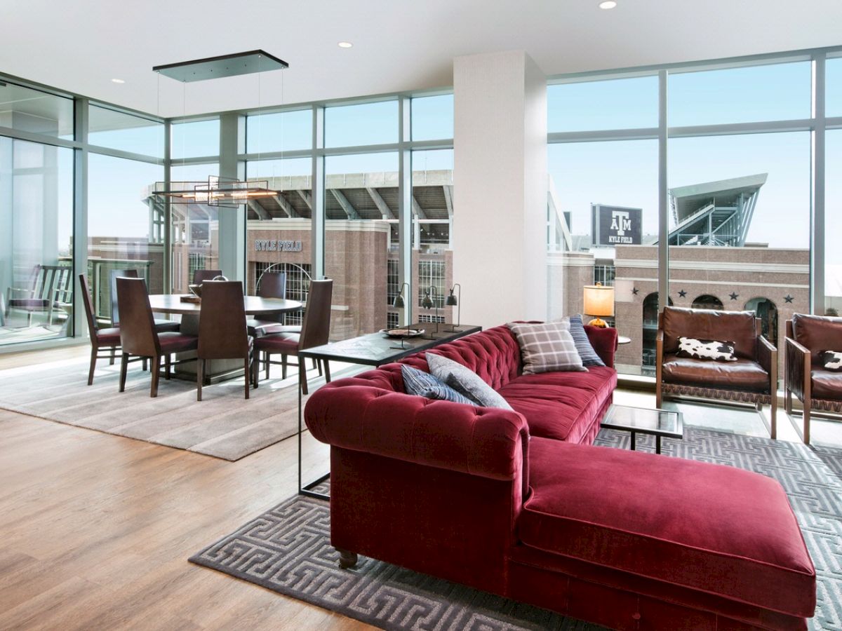 A modern hotel room with ceiling height windows overlooking Kyle Field featuring a maroon couch, dining area, and casual seating.