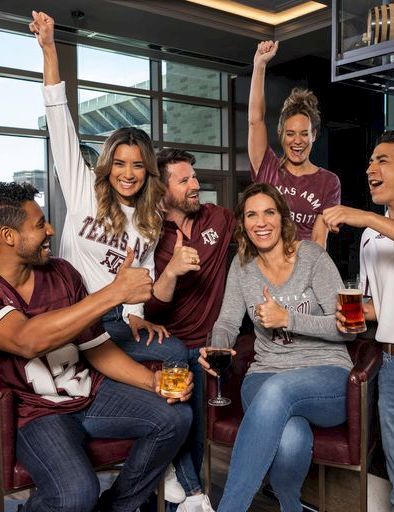 Group of friends at a sports bar, cheering with drinks, some wearing Texas gear, raising arms in excitement.