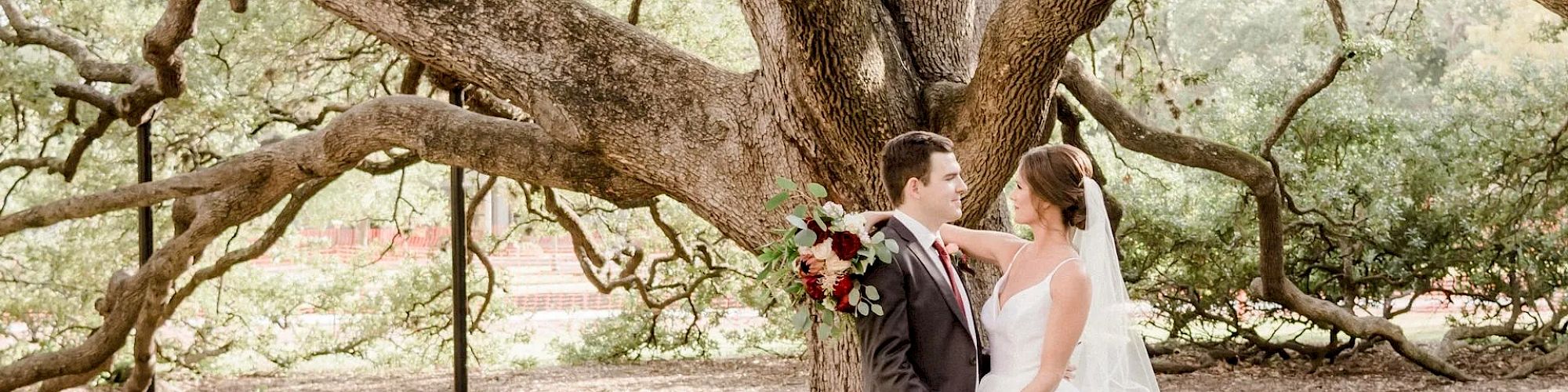 A bride and groom stand under a large, sprawling tree, holding hands and looking at each other, with the bride holding a bouquet of flowers.