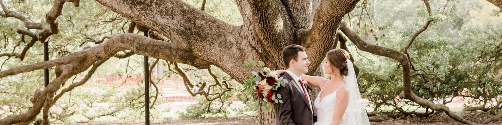 A couple dressed in wedding attire stands in front of a large, sprawling tree with intertwining branches, sharing an intimate moment.