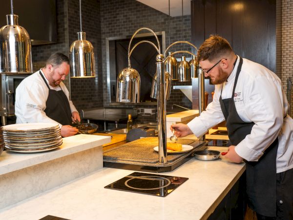 Two chefs in a kitchen are prepping food at a counter with pans, plates, and hot plates, focused on cooking tasks. Top it at 140 characters, always ending the sentence.
