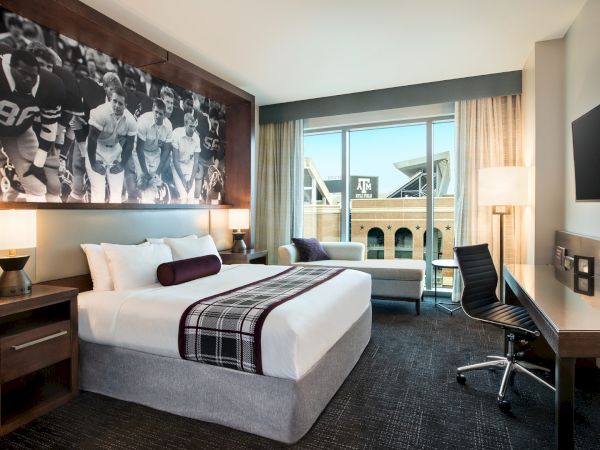 A modern hotel room with a large bed, wall art featuring a sports team, a desk, chair, and a large window with a view of a building.