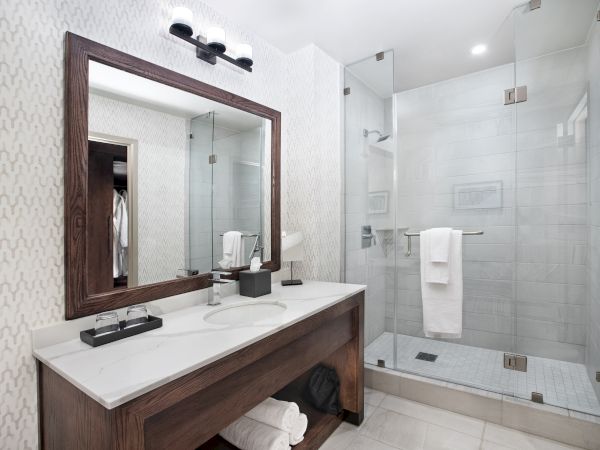 The image shows a modern bathroom with a wooden vanity, large mirror, and a glass-enclosed shower. The space is clean and well-lit.