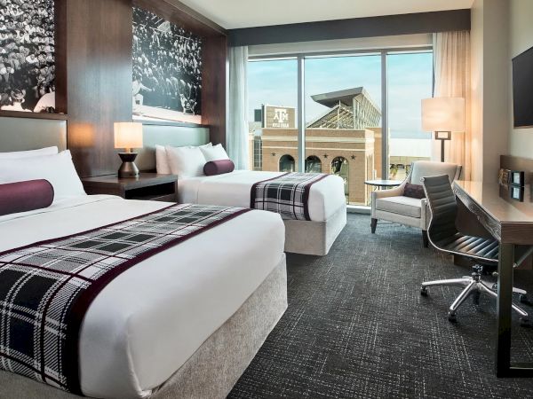 Modern hotel room with two queen beds, a desk and office chair, lounging chair, and a window overlooking Kyle Field