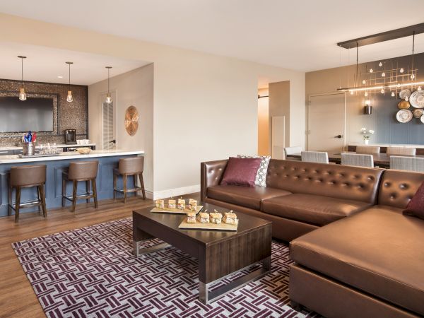 Modern hospitality suite with a brown leather couch, barstools along the kitchen area, a dining room table and eccentric wall decor
