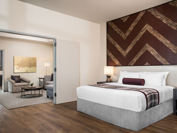 A modern hotel room with a large bed, decorative headboard, and shared living area featuring couches, a coffee table, and a piece of artwork on the wall.