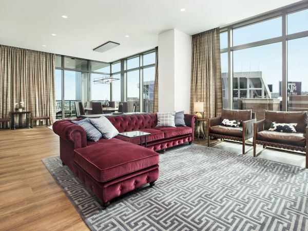 A modern penthouse suite with a maroon couch, grey carpet, leather chairs, dining area, and ceiling height windows