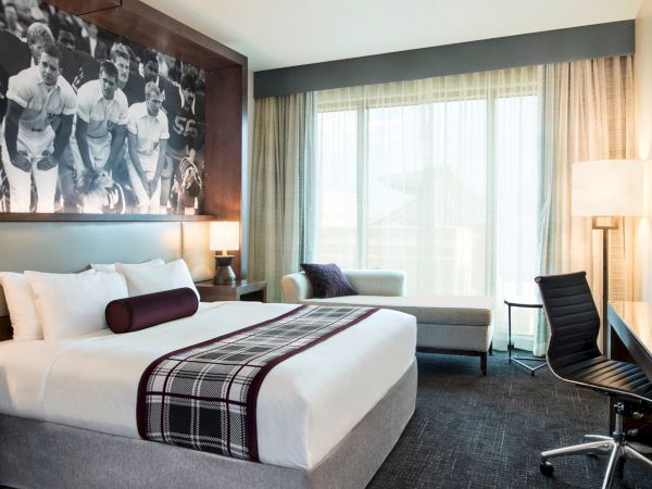 A modern hotel room with a large bed, a desk with a chair, a sofa, and a large window. A black and white sports-themed mural is on the wall.