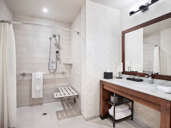 The image shows a modern bathroom with a walk-in shower, a bench, a large mirror, a sink with countertop, and various toiletries.