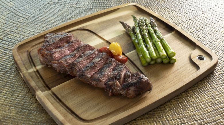 A wooden plate with a grilled steak and asparagus spears on a textured surface, garnished with a small bell pepper and a clove of garlic.