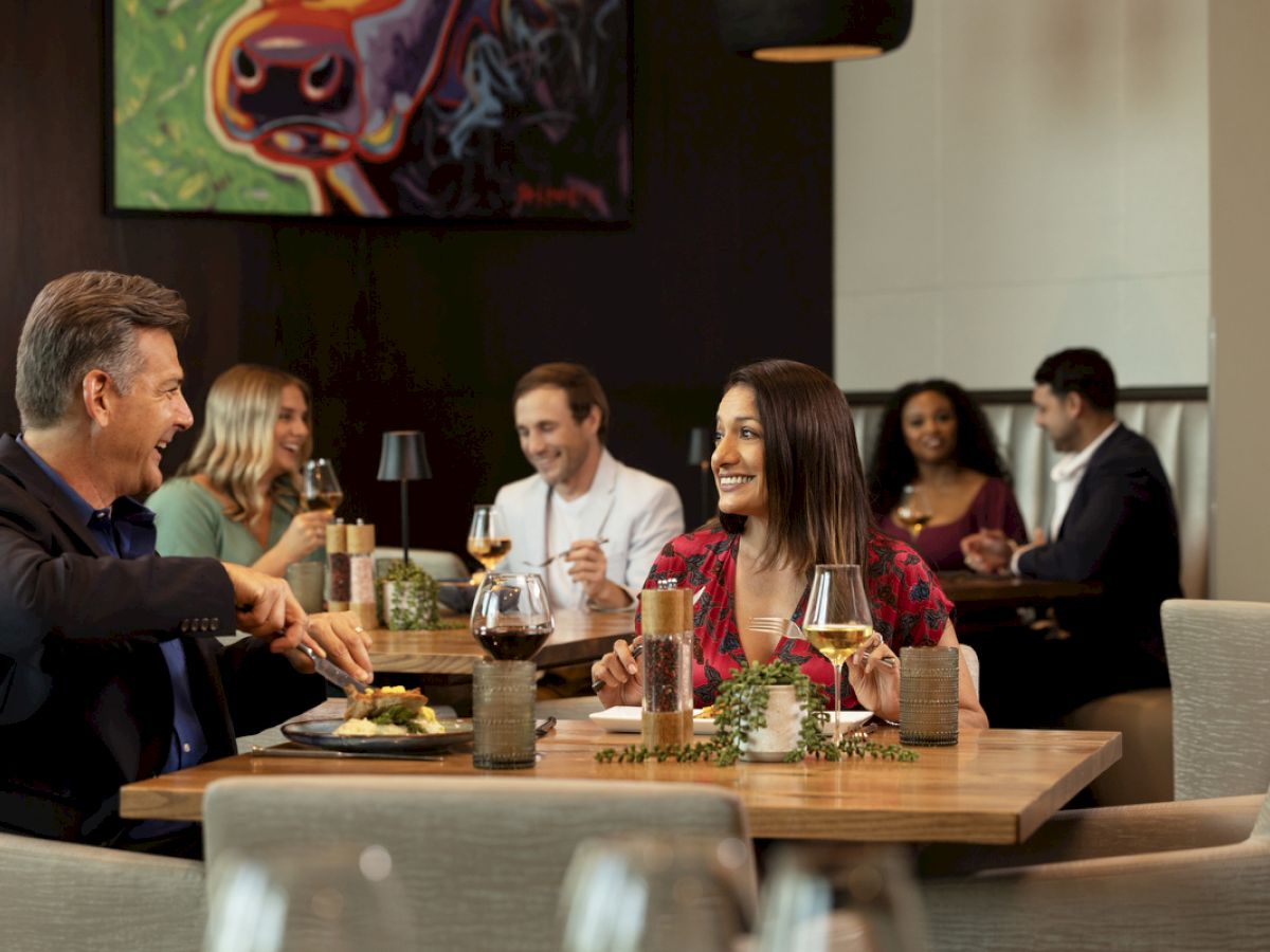 Two couples are dining in Brazos - PTK, enjoying their meals and conversations. The ambiance is cozy with modern decor and a colorful painting.