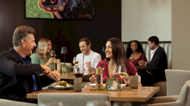 Two couples are dining in Brazos - PTK, enjoying their meals and conversations. The ambiance is cozy with modern decor and a colorful painting.