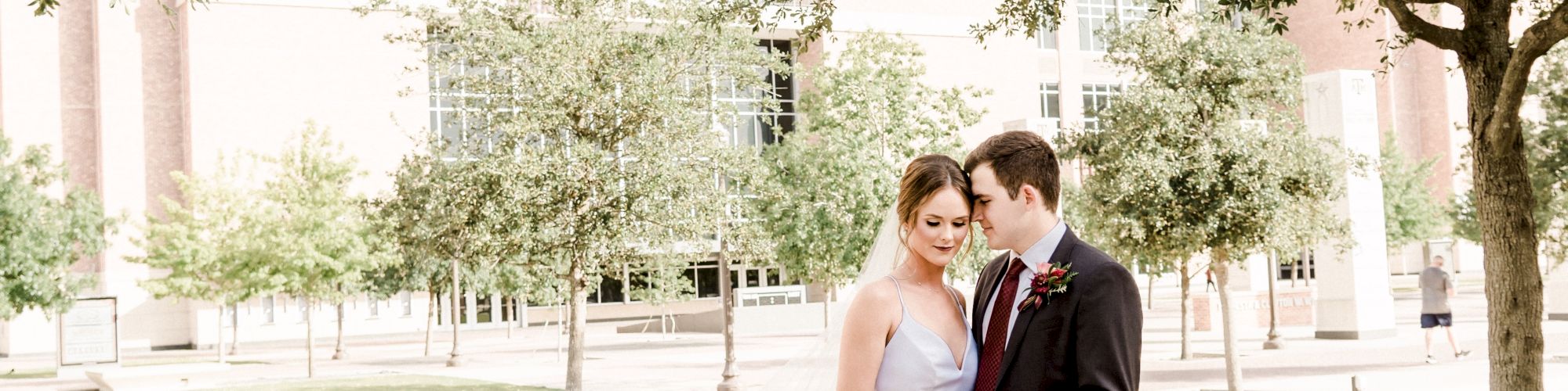 A couple in wedding attire is standing close, outdoors, near a large building with trees around them, looking at each other lovingly.