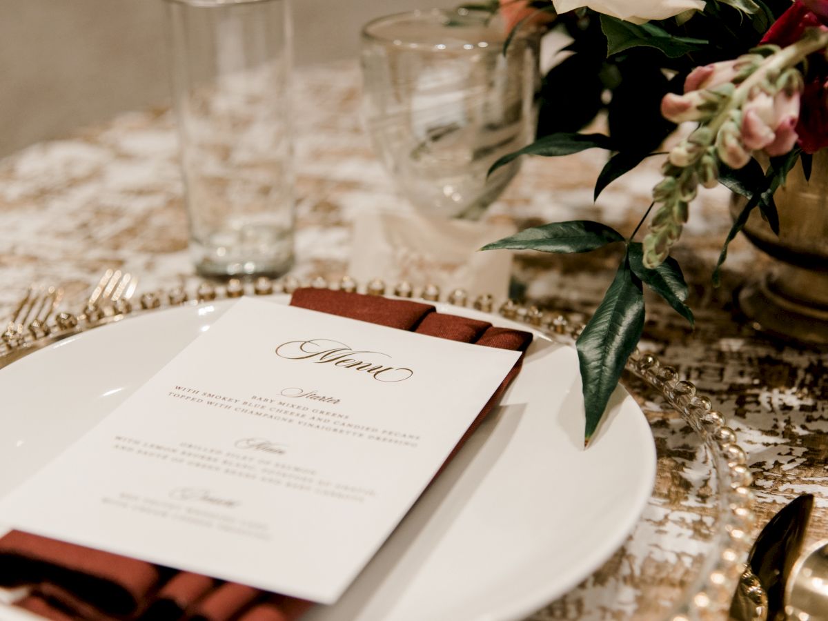 A decorated dining table with a menu on a plate, accompanied by a red napkin, and surrounded by glassware, cutlery, and floral arrangements.