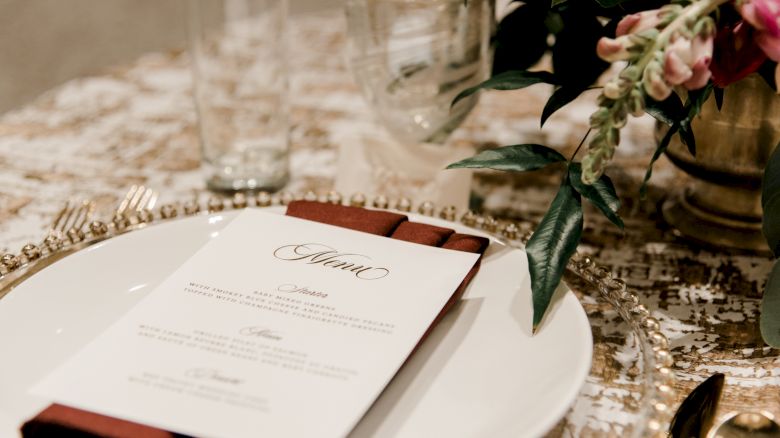 A decorated dining table with a menu on a plate, accompanied by a red napkin, and surrounded by glassware, cutlery, and floral arrangements.