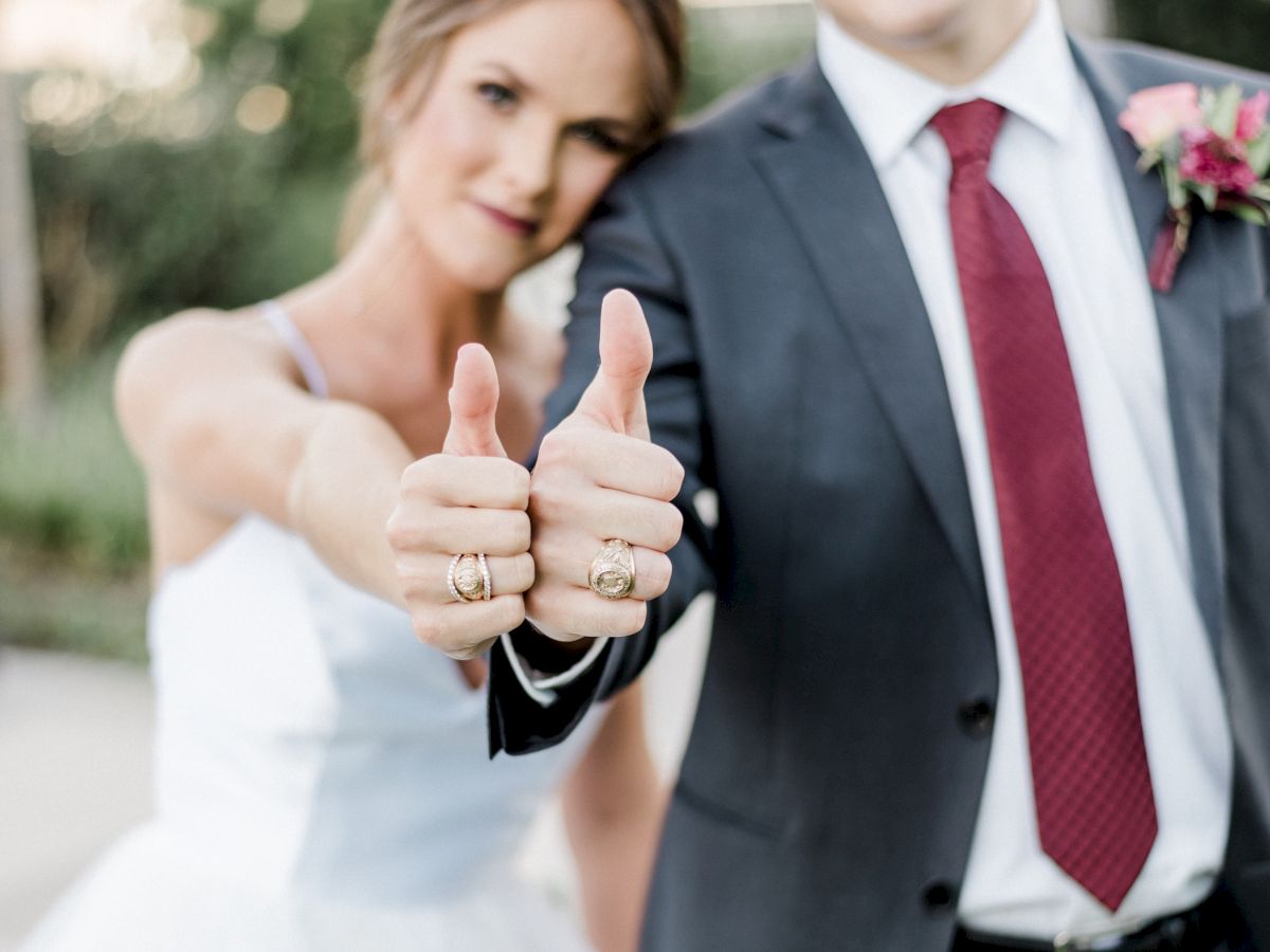 A couple is dressed in wedding attire, both giving thumbs up and appearing happy together in an outdoor setting, with greenery in the background.
