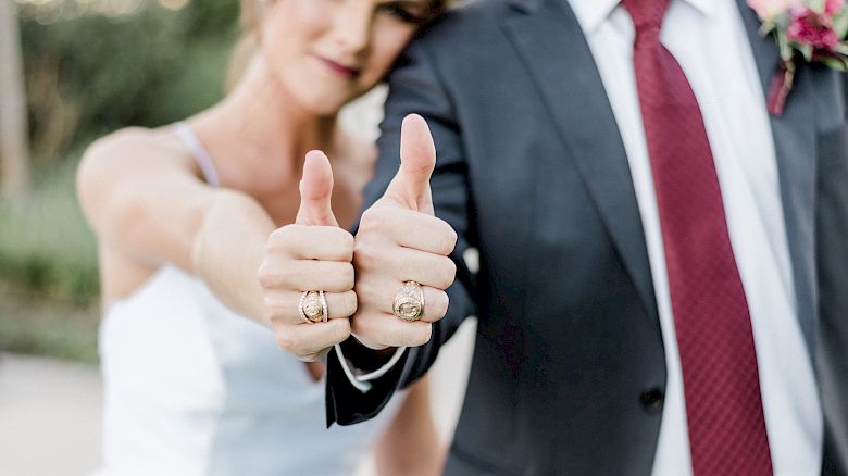 A couple is dressed in wedding attire, both giving thumbs up and appearing happy together in an outdoor setting, with greenery in the background.