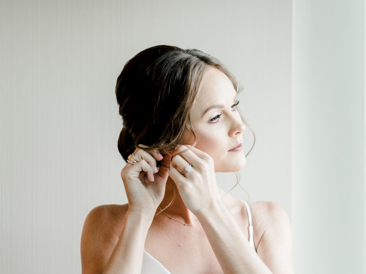A bride in a white dress is adjusting her earring, looking off to the side in a softly lit room.