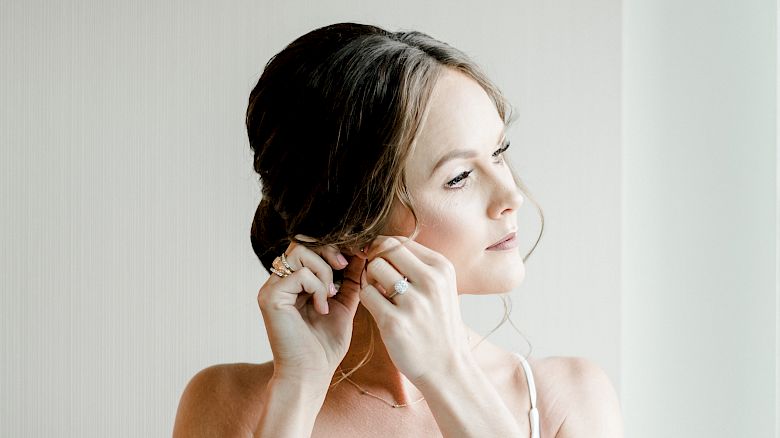 A bride in a white dress is adjusting her earring, looking off to the side in a softly lit room.