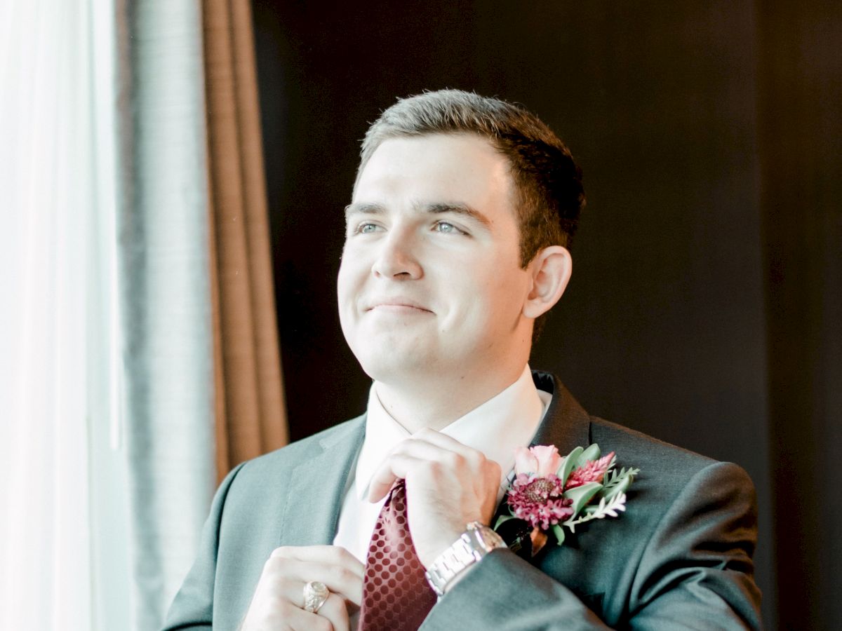 A man in a suit adjusts his tie and stands near a window. He has a floral boutonniere on his jacket.
