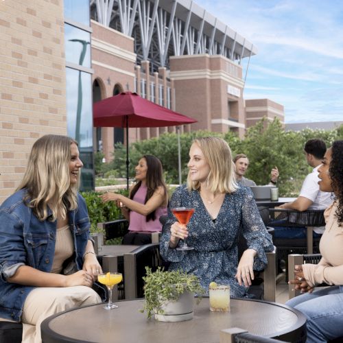 Group of friends chatting and sipping drinks on a sunny patio at a mall or venue, with umbrellas, comfy chairs, and table plants.