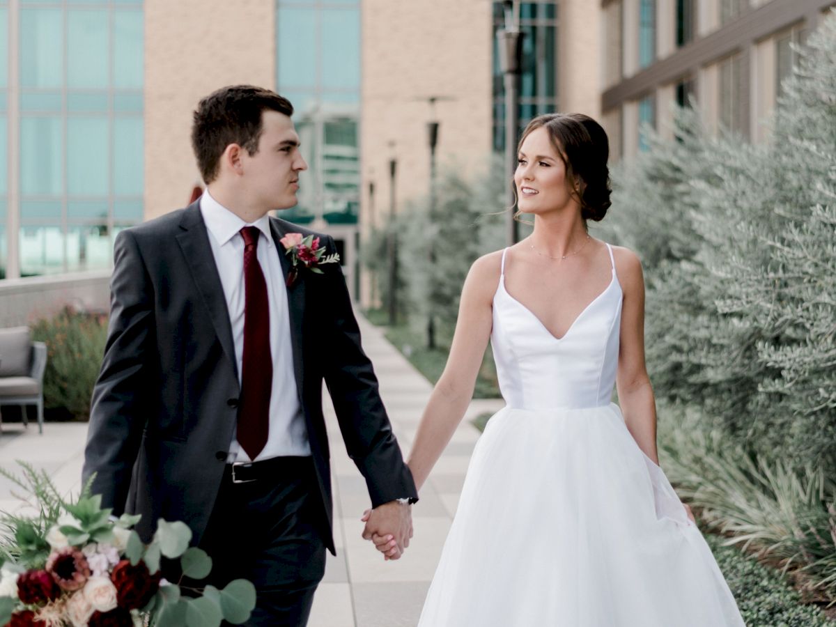 A couple dressed in wedding attire is walking hand in hand outdoors, surrounded by greenery and a building.