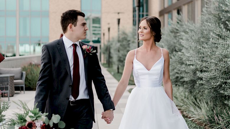 A couple dressed in wedding attire is walking hand in hand outdoors, surrounded by greenery and a building.