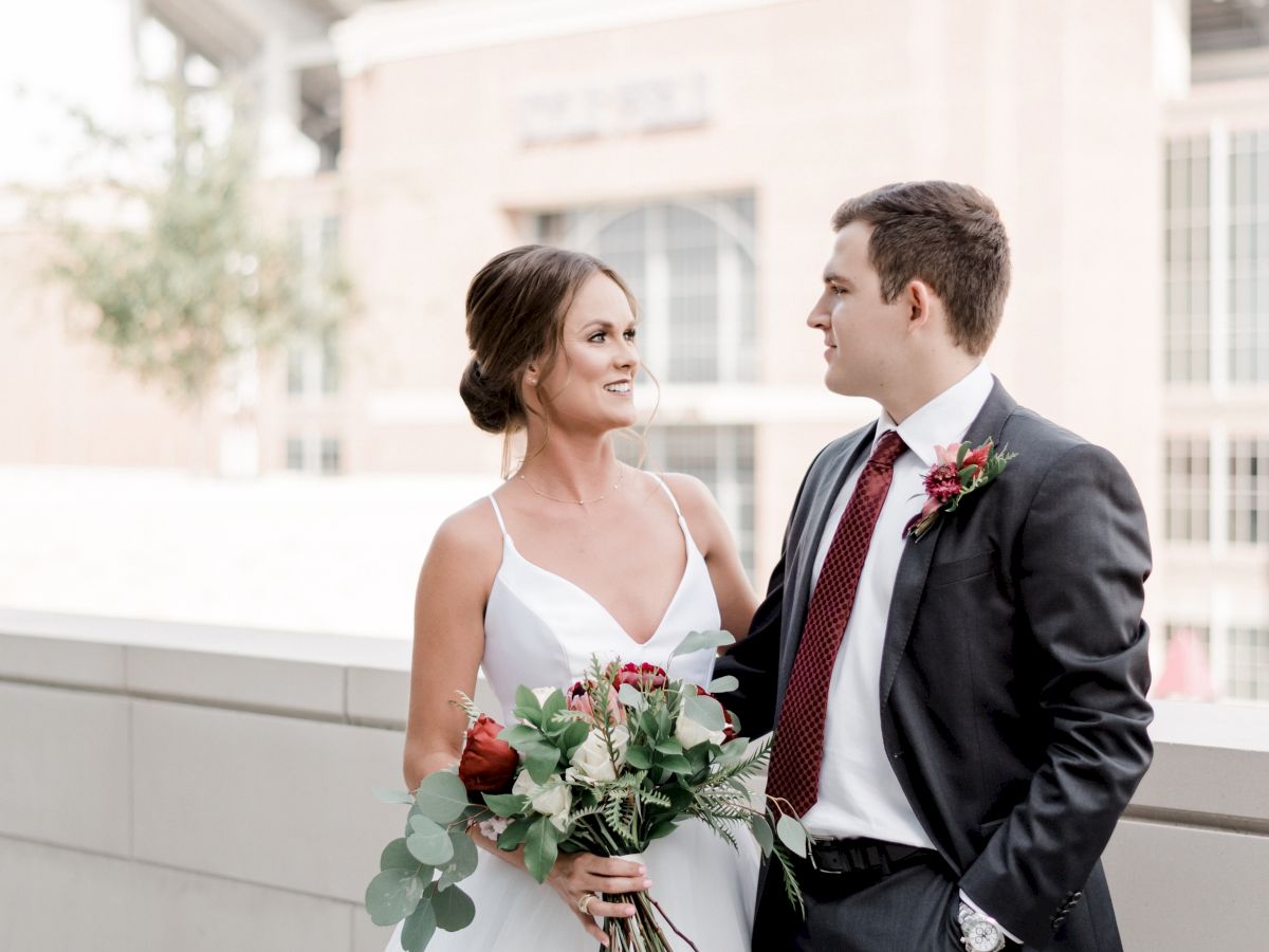 A bride and groom are standing together outdoors, with the bride holding a bouquet, and a stadium is visible in the background.
