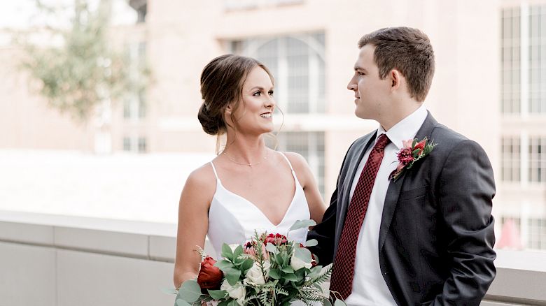 A bride and groom are standing together outdoors, with the bride holding a bouquet, and a stadium is visible in the background.