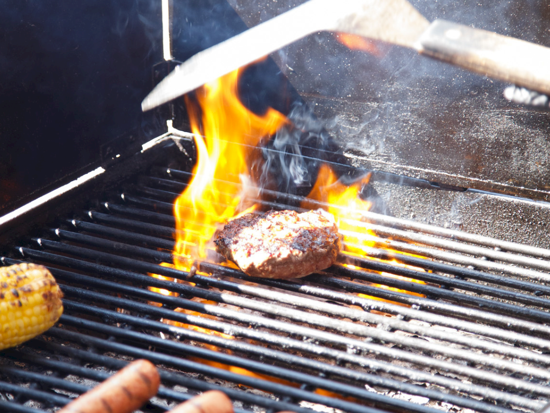 Grilling on a charcoal barbecue: flames lick a steak as tongs flip it, with sausages and corn nearby on the grates.