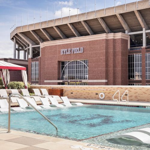 An outdoor pool area beside a brick stadium, with lounge chairs, red umbrellas, and a building labeled &ldquo;METRO&rdquo; in the background.