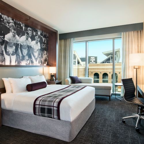 A modern hotel room with a large bed, black-and-white mural, plaid throw, two nightstands, a desk and chair, a seating area by the window, and warm lighting.
