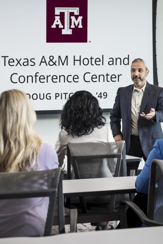 A person is giving a presentation to a small group at the Texas A&M Hotel and Conference Center, as shown on the screen behind him.