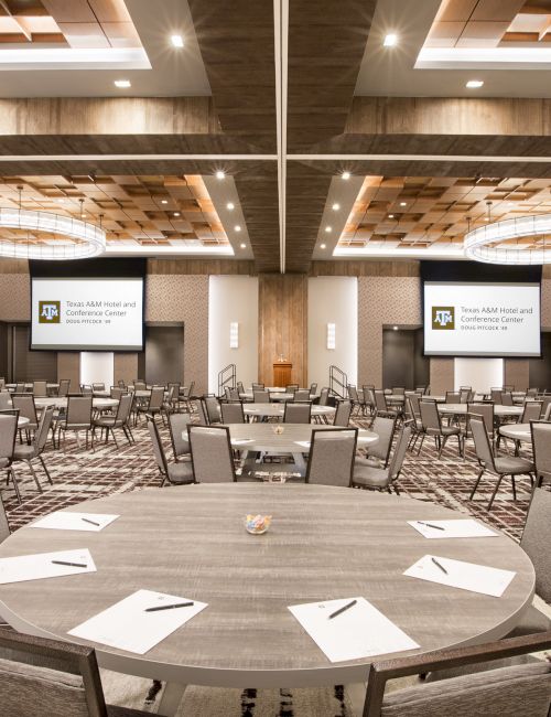 A spacious conference room with circular tables and chairs, each table set with notepads and papers. Two large screens display a presentation. Ending the sentence.