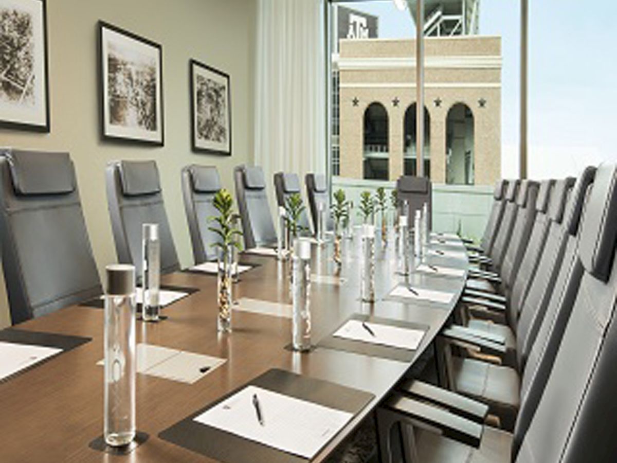 A modern conference room with a long table, chairs, water bottles, and notepads, facing large windows overlooking a building outside.